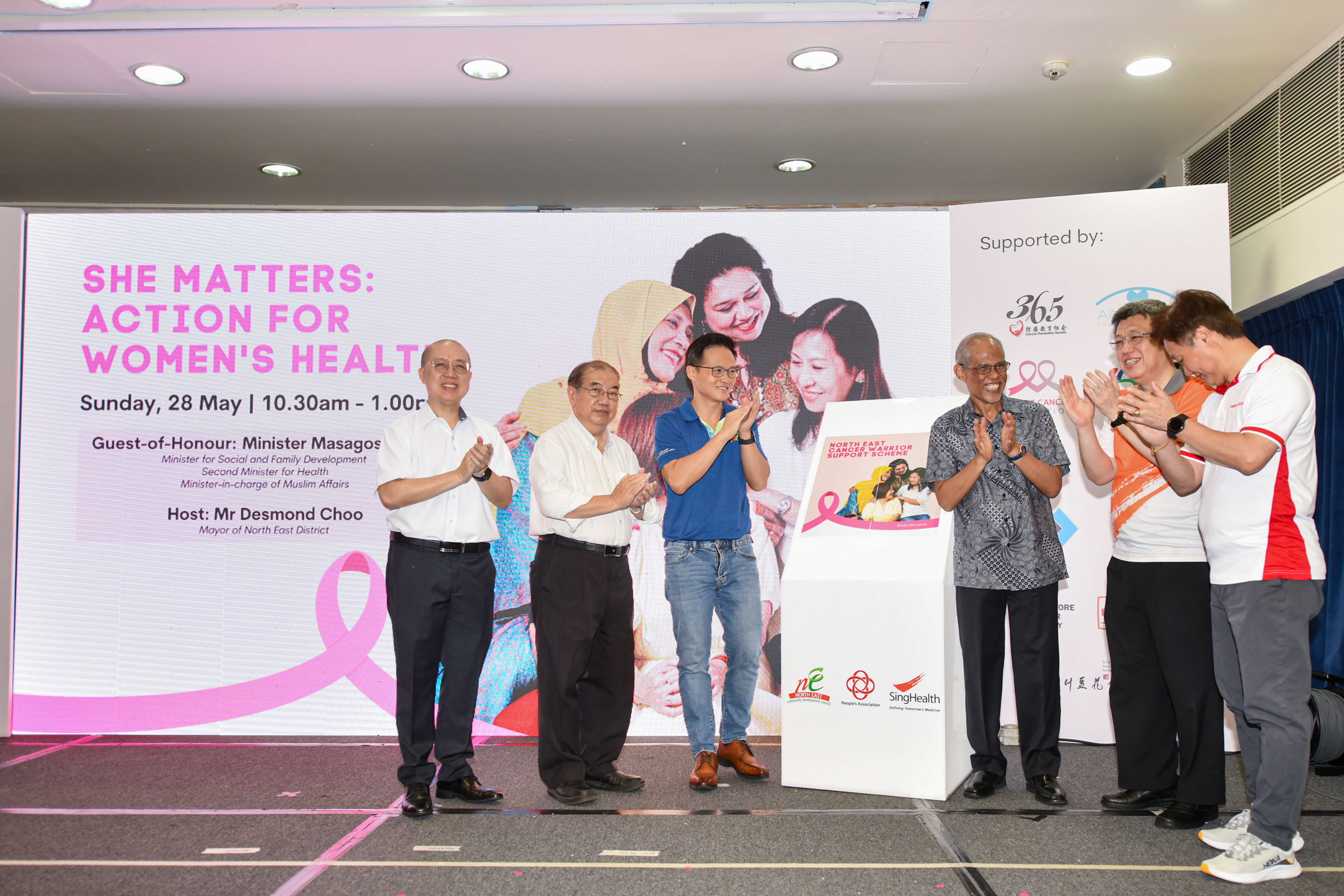 Several men stand on a stage, clapping in front of a banner promoting women's health awareness.